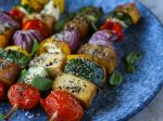 Barevné grilované špízy se zeleninou, kostkami tofu a čerstvými bylinkami na modrém talíři.
